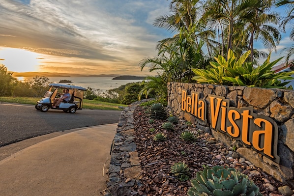 A golf cart parked near a sign reading "Bella Vista" with a stone wall and tropical plants. - Bella Vista East 5 (Whitsundays)