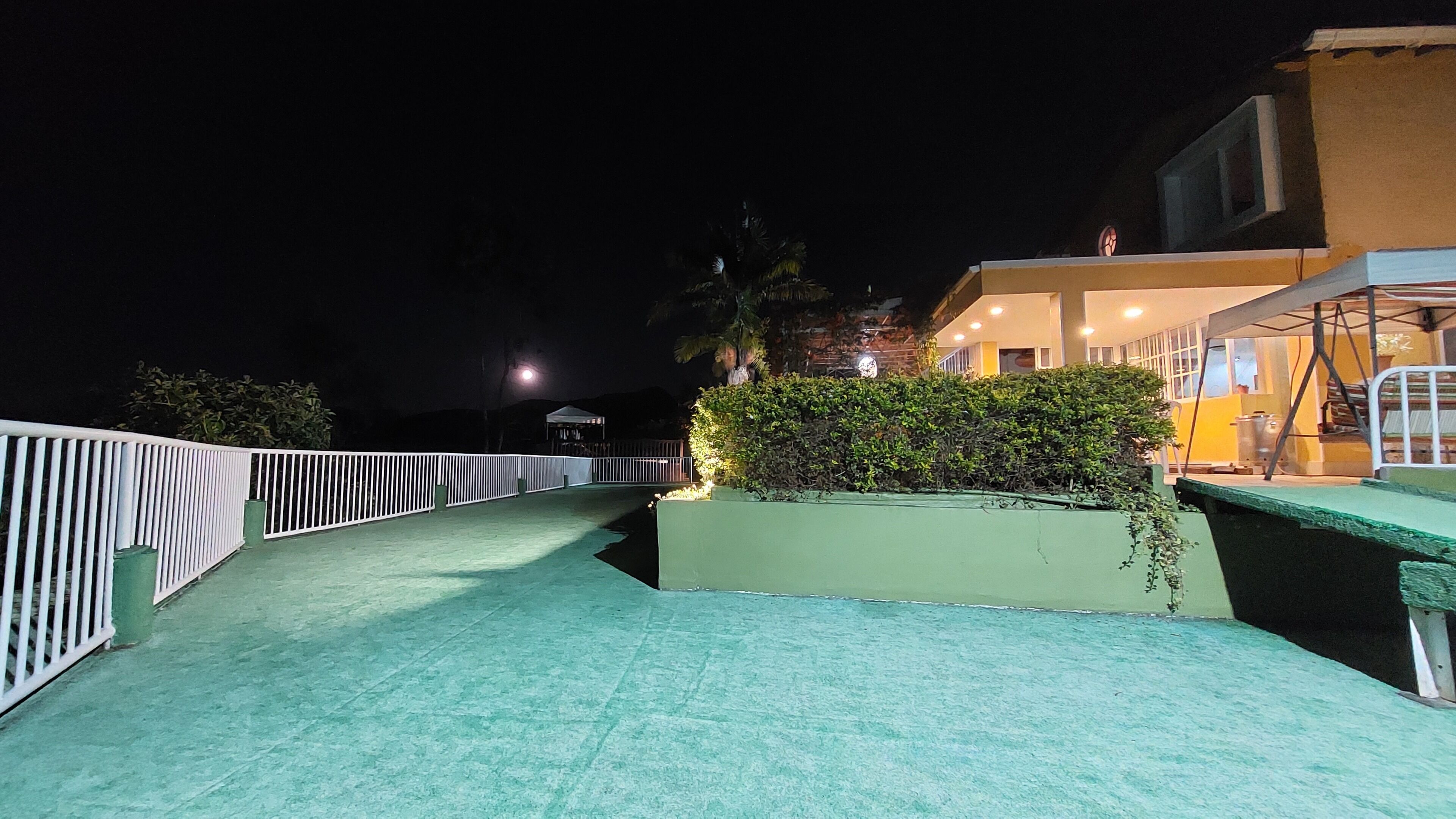 A nighttime view of a rooftop terrace with a white railing, a green planter with shrubs, and a building with illuminated windows in the background.