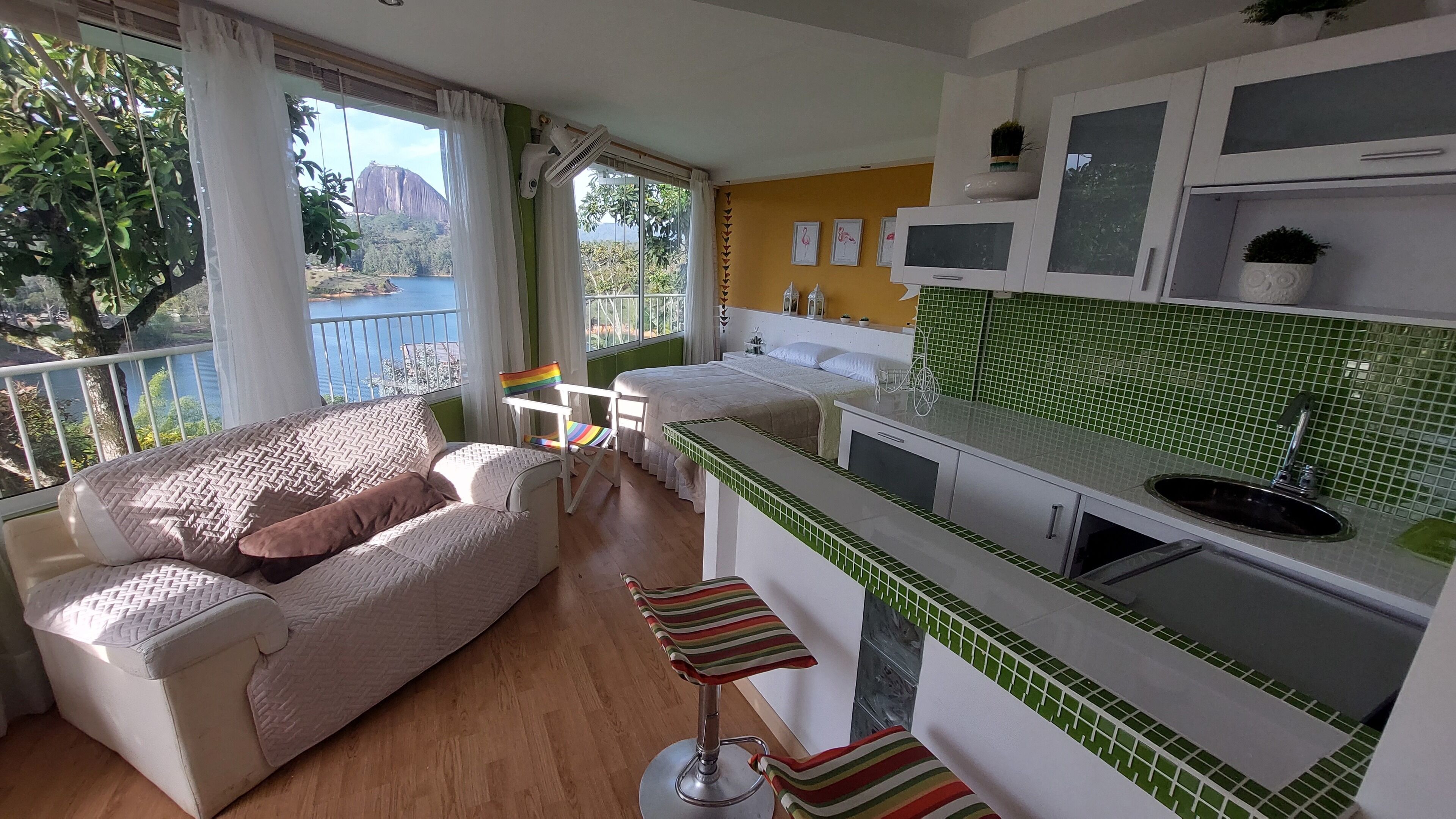 A modern kitchen with a green tiled backsplash, a white countertop, and a sink. There is a dining area with a white table and chairs, and a balcony with a view of a mountain and a lake.