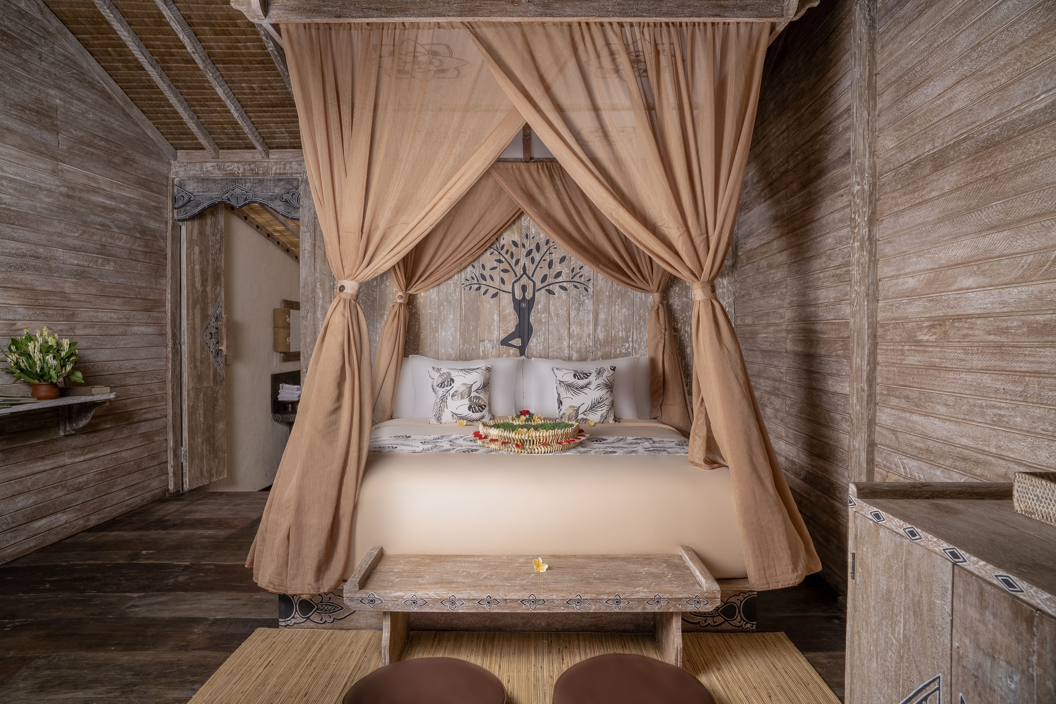 A room with a bed under a canopy, wooden walls, a small table, and a decorative tree silhouette on the wall.