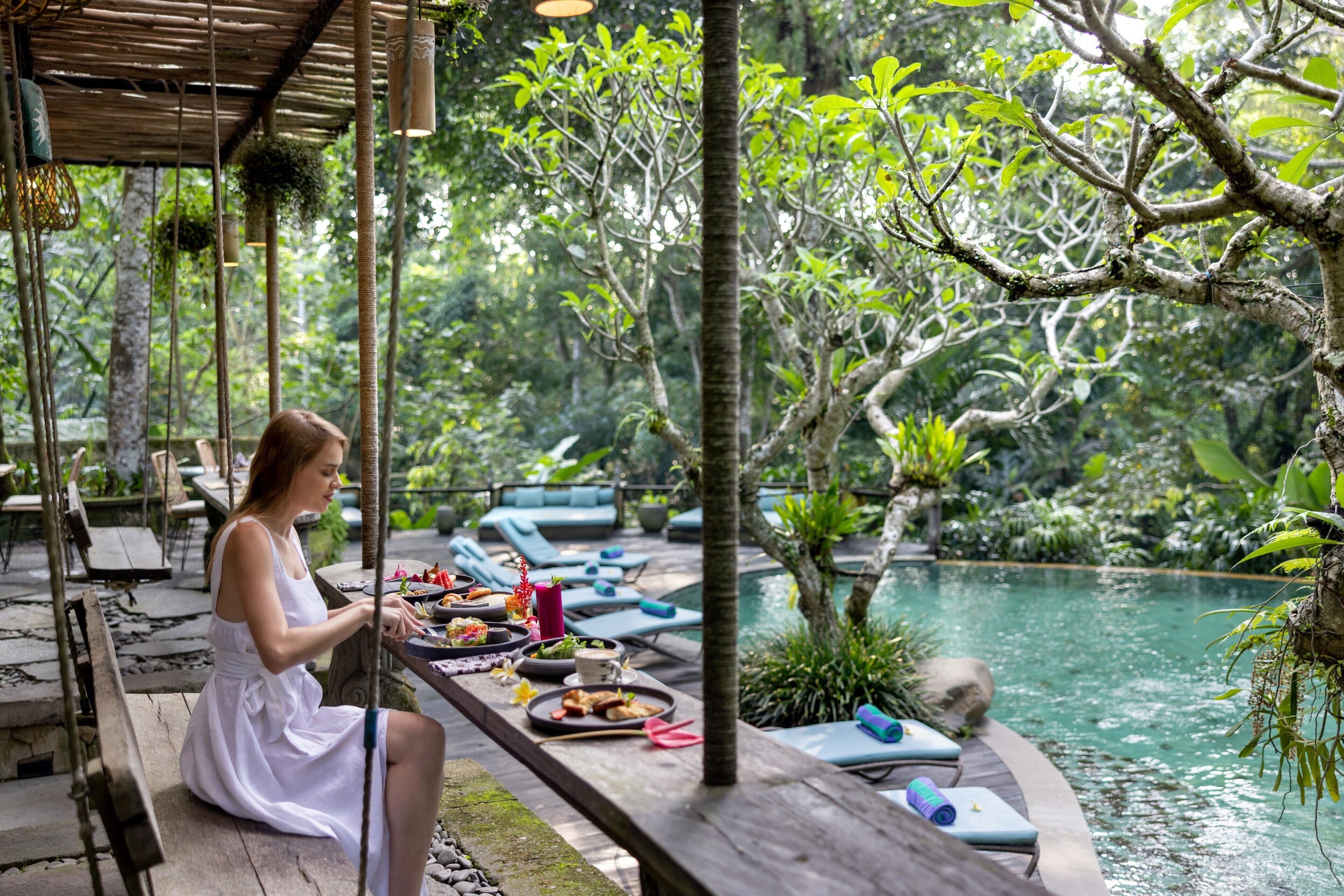 A woman sitting at a wooden table with food and drinks, overlooking a pool and lush greenery.