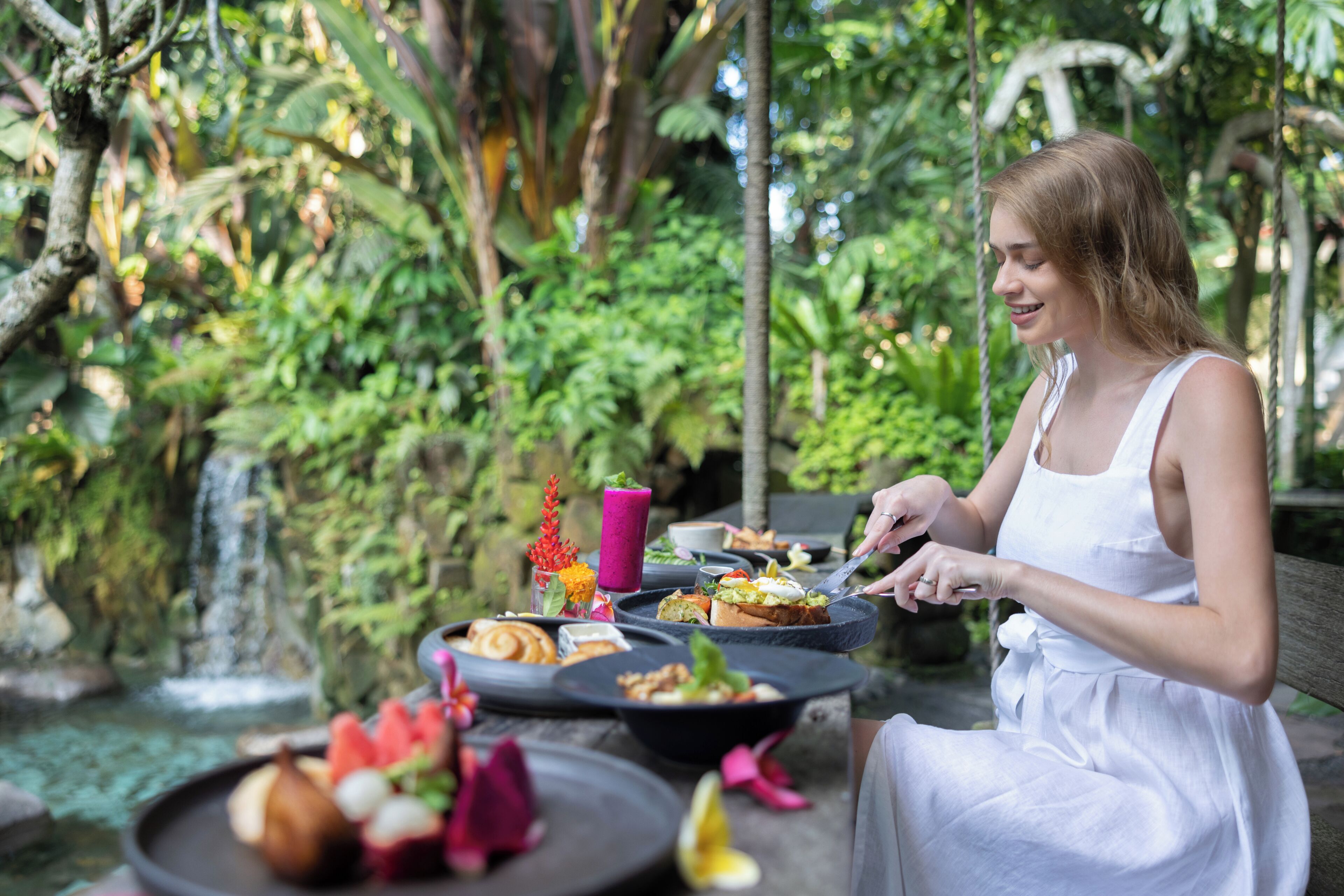 A person dining outdoors with a variety of food items on a tray, surrounded by lush greenery and a waterfall in the background.