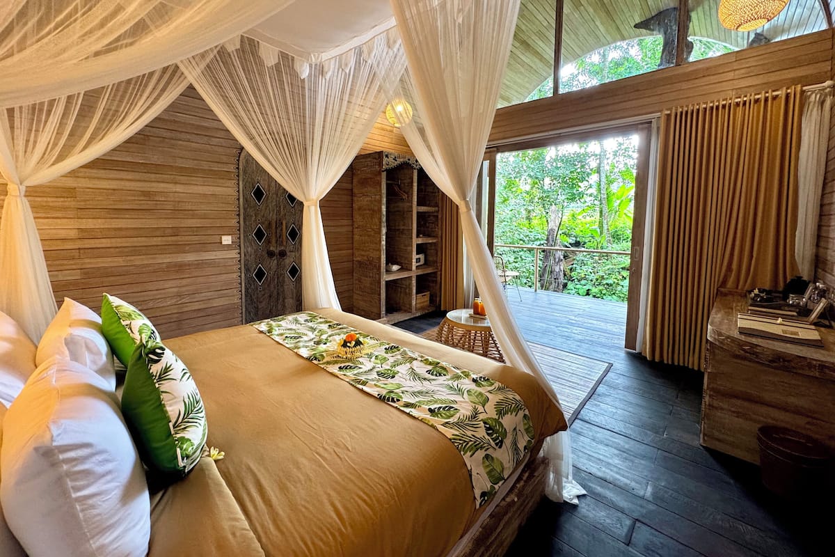 Une chambre à coucher avec un lit à baldaquin, des murs en bois, une petite table avec une boisson, et une vue sur la verdure à l’extérieur.
