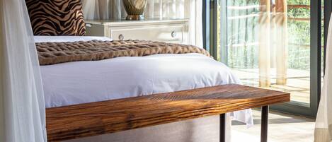 A bedroom with a canopy bed, a wooden bench, a nightstand with a lamp, and a view of greenery outside.