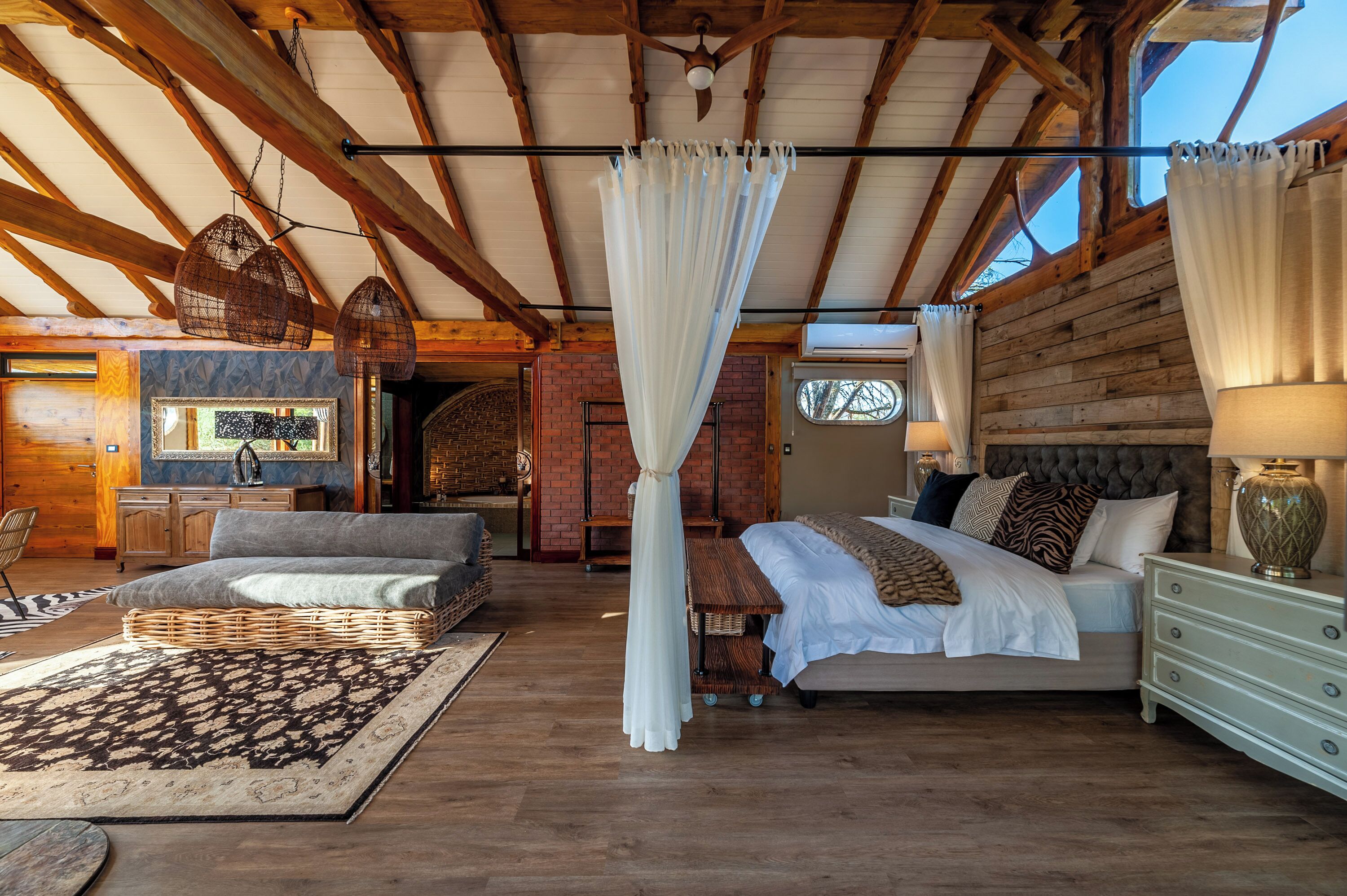 A spacious bedroom with a large bed, a sitting area with a sofa and a small table, and a view of a brick wall and wooden ceiling beams.