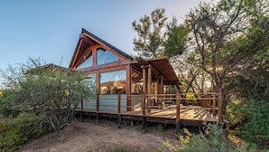 A wooden cabin with a large glass window, surrounded by trees and a dirt path. - Mont Eco Game Reserve (Montagu)