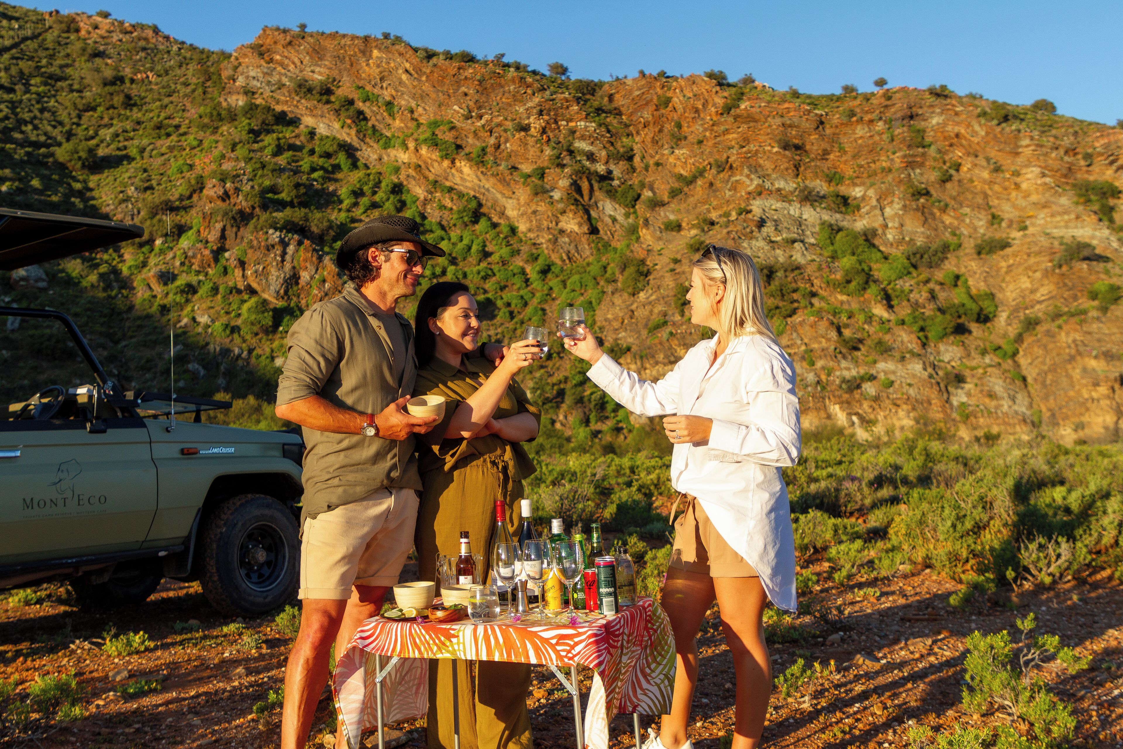 A group of people toasting with drinks around a table with bottles and glasses, a car, and a hilly landscape in the background.