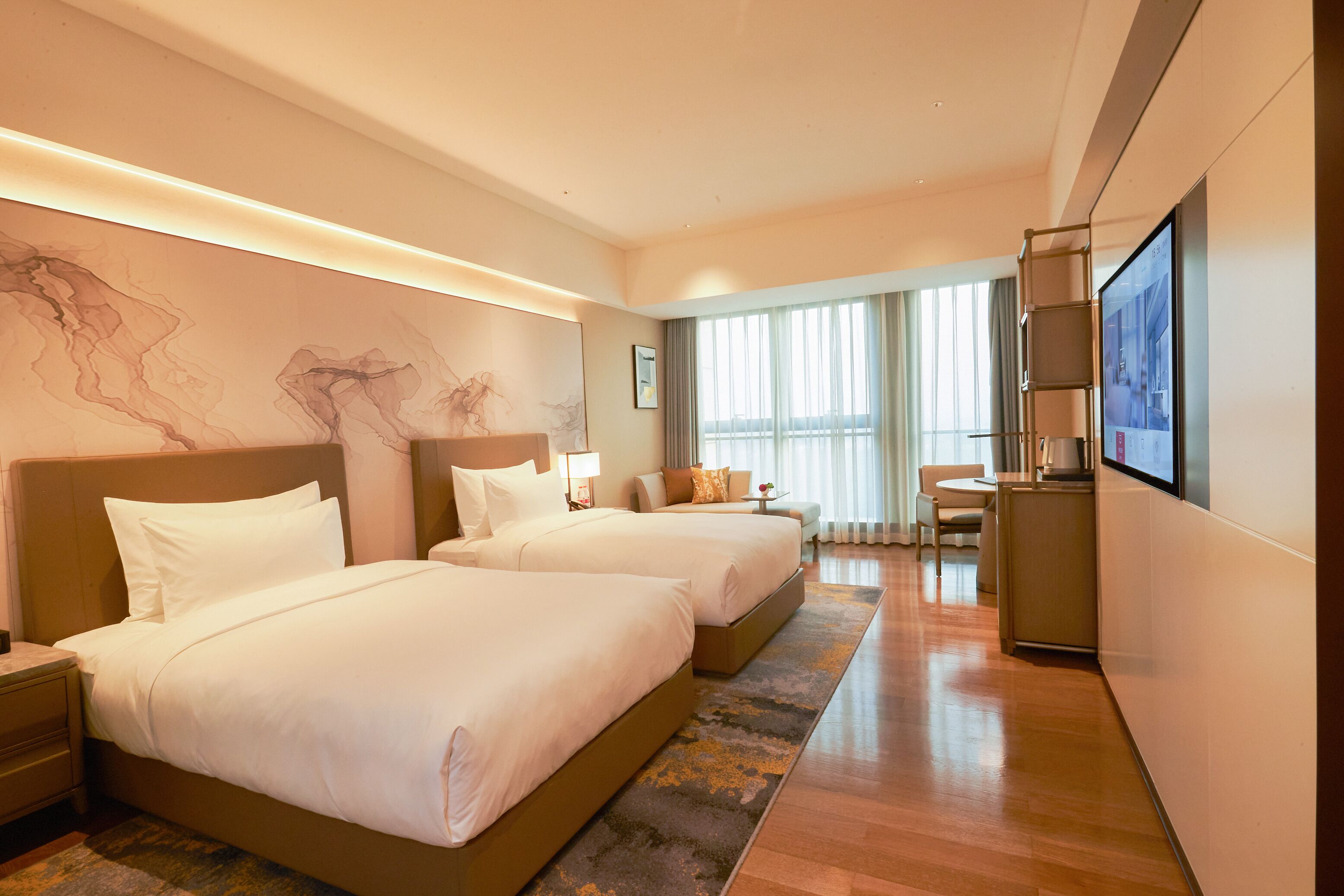 A hotel room with two beds, a desk, a chair, a TV, and a large window with curtains.