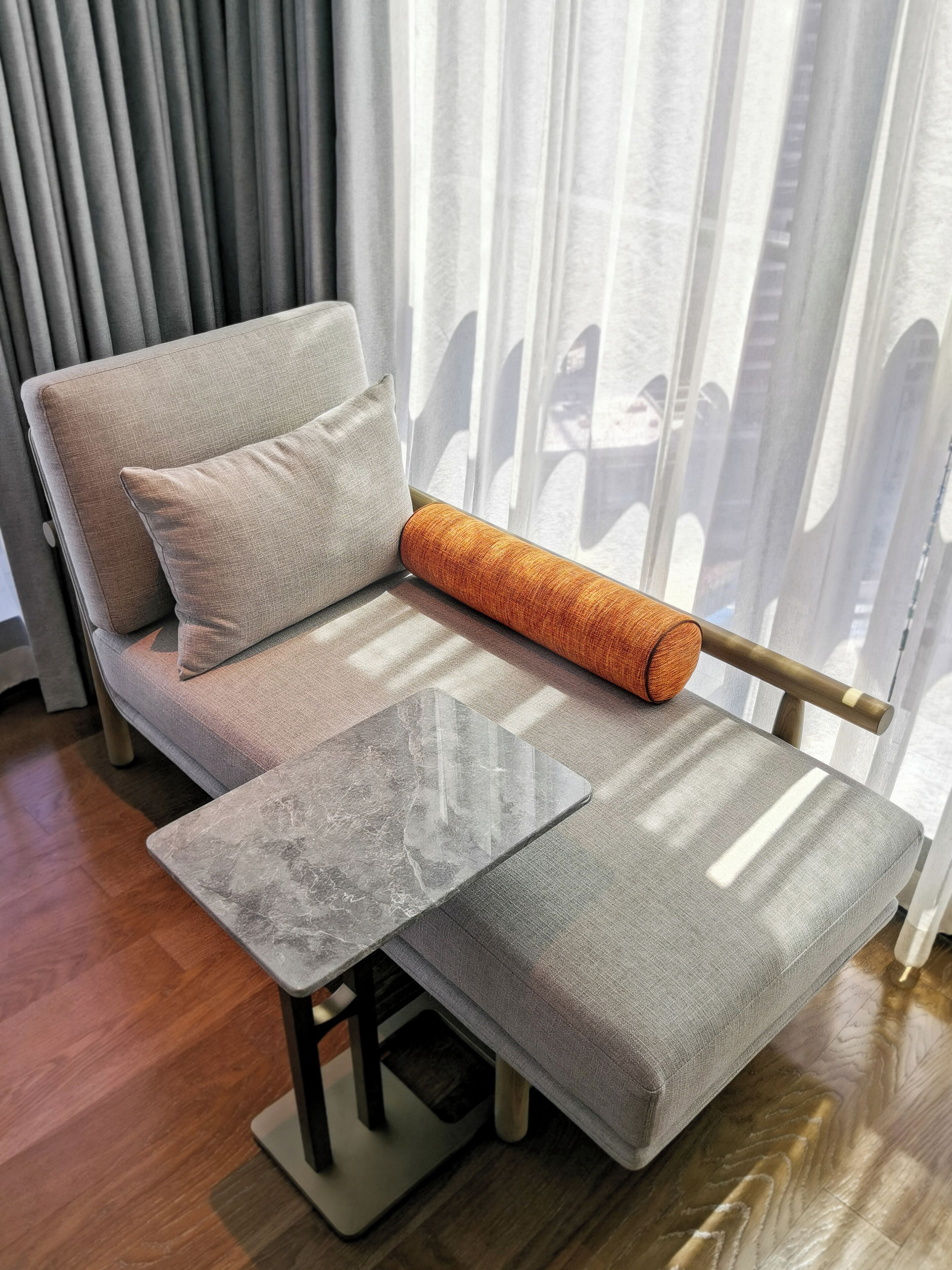Un sofá moderno con una mesita de madera y un cojín, ubicado frente a una ventana con cortinas translúcidas.
