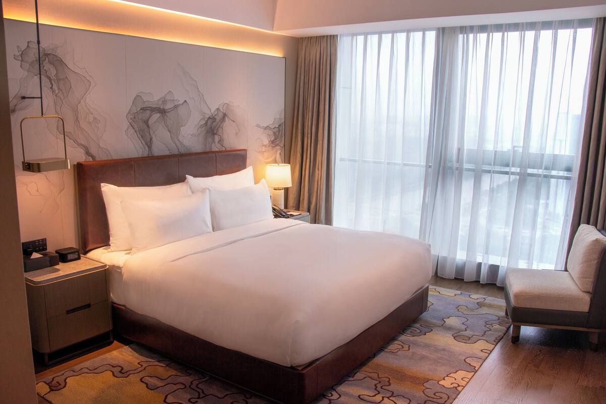 Een hotelkamer met een groot bed, een nachtkastje met een lamp, een stoel en uitzicht op het buitenleven door dunne gordijnen.