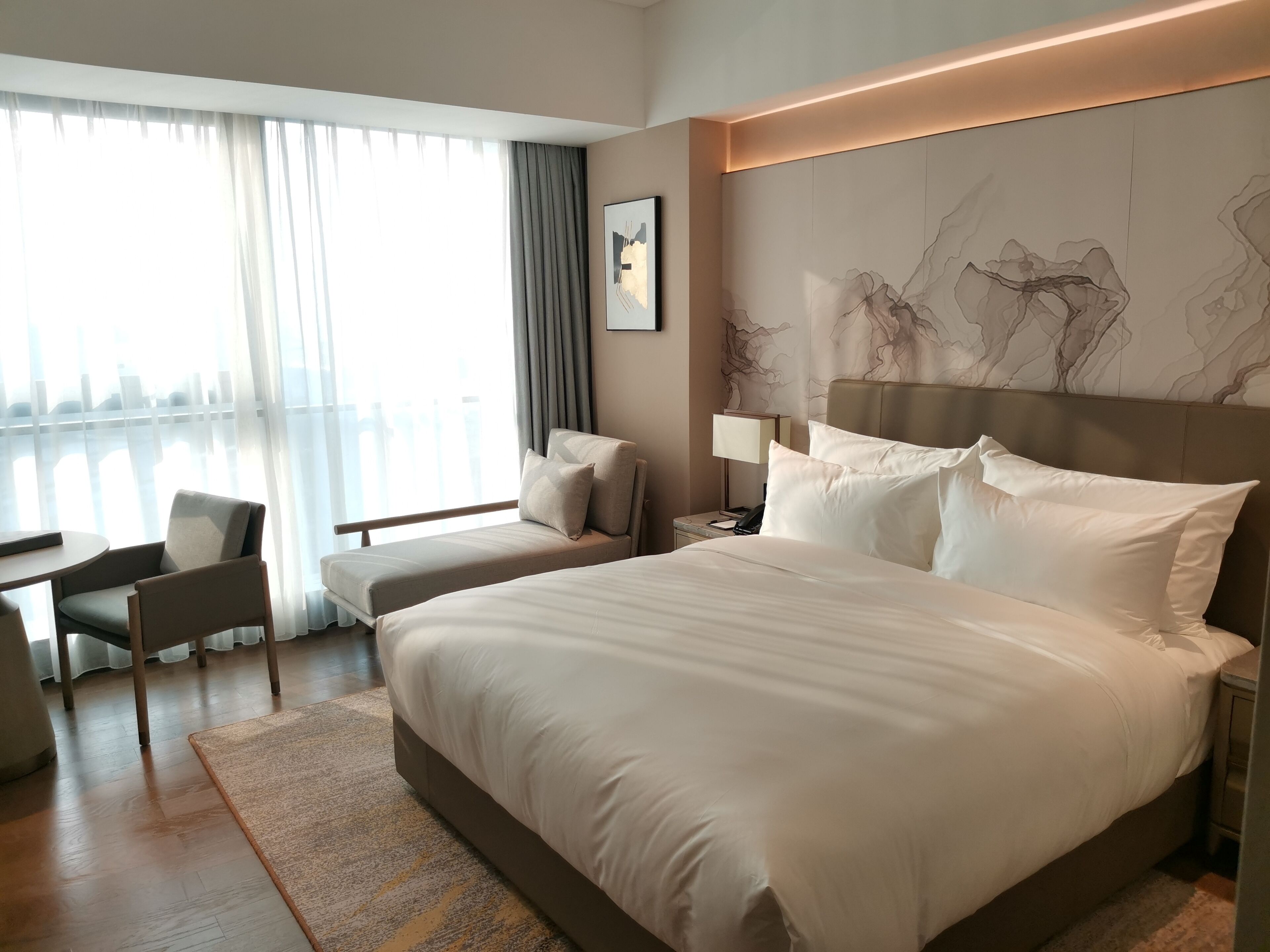 Une chambre d’hôtel avec un grand lit, un bureau et une chaise. Il y a une fenêtre avec des rideaux, une lampe fixée au mur et un tableau encadré.
