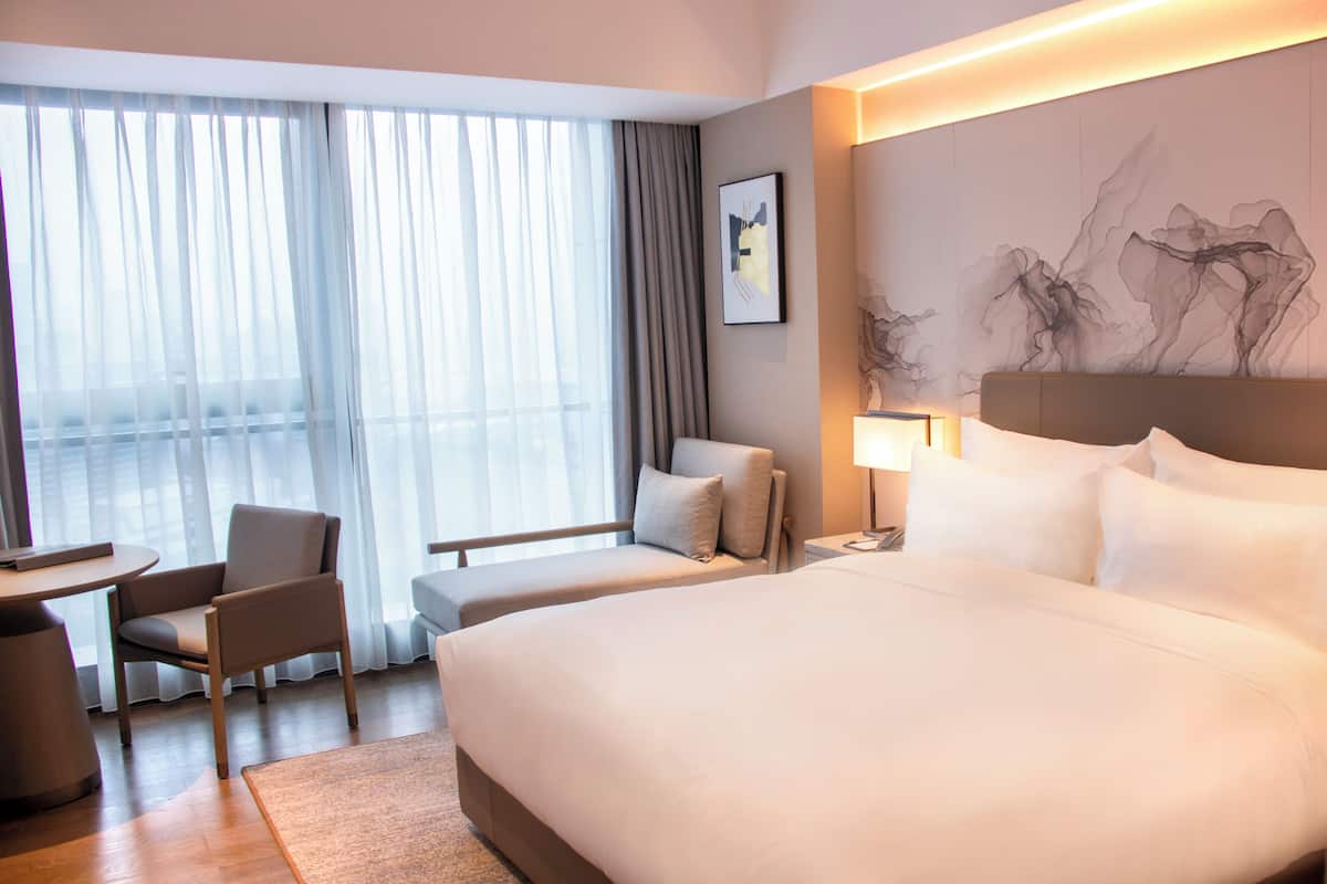 Een hotelkamer met een groot bed, een bureau met een stoel, een kleine tafel en een groot raam met dunne gordijnen.