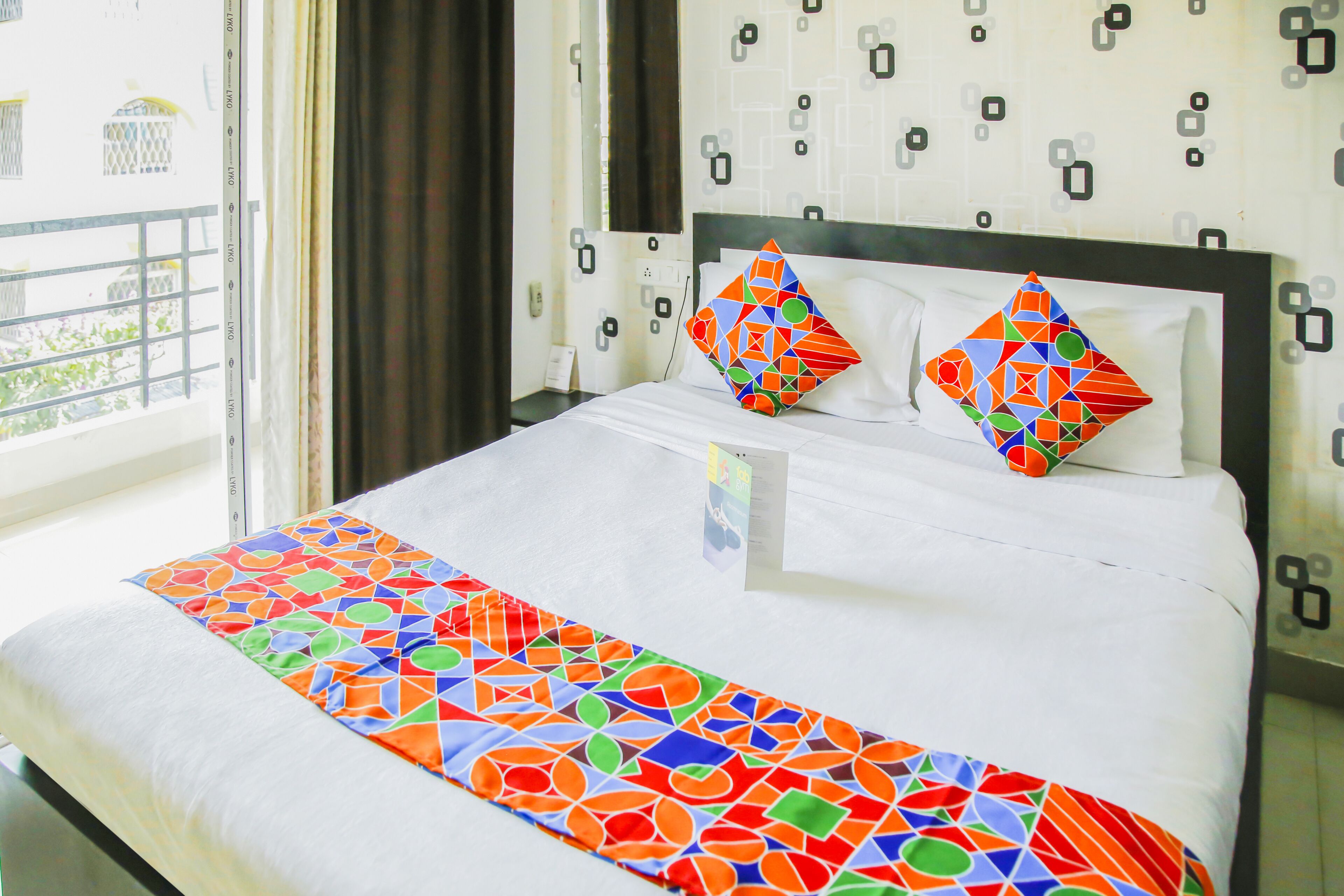A hotel room with a bed, two pillows with geometric patterns, a window with a view of greenery, and a patterned wall.