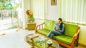 A green sofa with a person sitting on it, a glass coffee table with a cup and saucer, and a small side table with a picture frame and flowers. - FabHotel Eleanora Inn (Barasat)