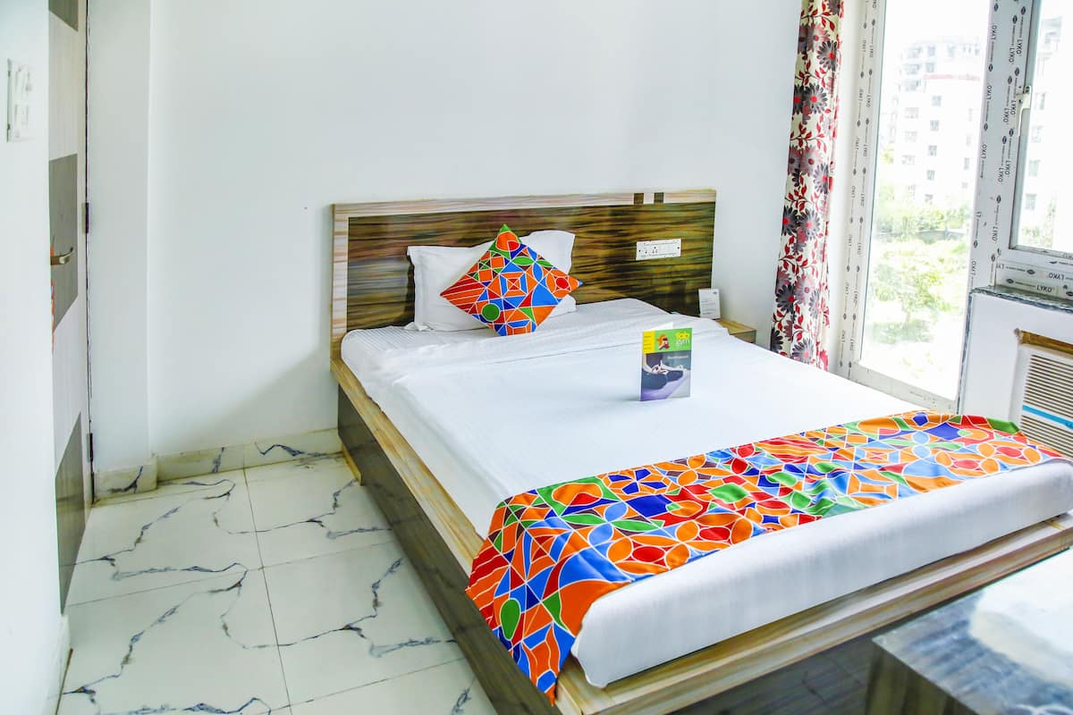 A hotel room with a wooden headboard, a bed with a colorful geometric-patterned bedspread, a white pillow, and a window with a view of buildings and trees.