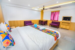 Una habitación de hotel con una cama grande, dos sillones morados, una mesita, un televisor y un ventilador de techo.