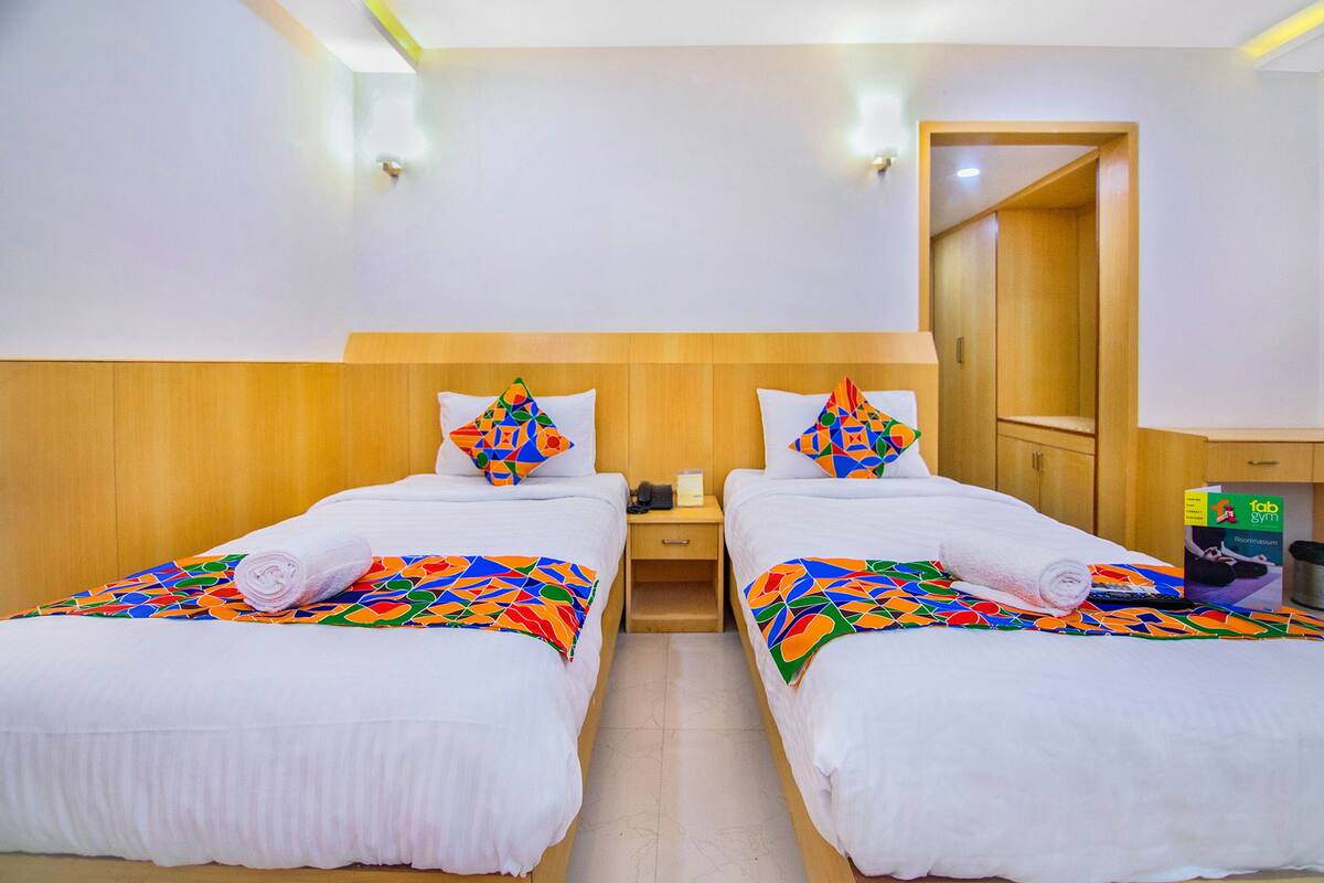 Hotelkamer met twee bedden, elk voorzien van een kleurrijk beddegoed met een geometrisch patroon en bijpassende kussens. Een houten hoofdbord, een nachtkastje met een telefoon en een doos, en een spiegel aan de muur.