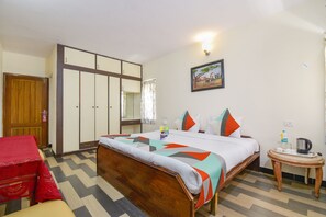A hotel room with a bed, a wooden wardrobe, a small table with a kettle, and a red chair. - Fabexpress Wenlock Homes (Ootacamund)