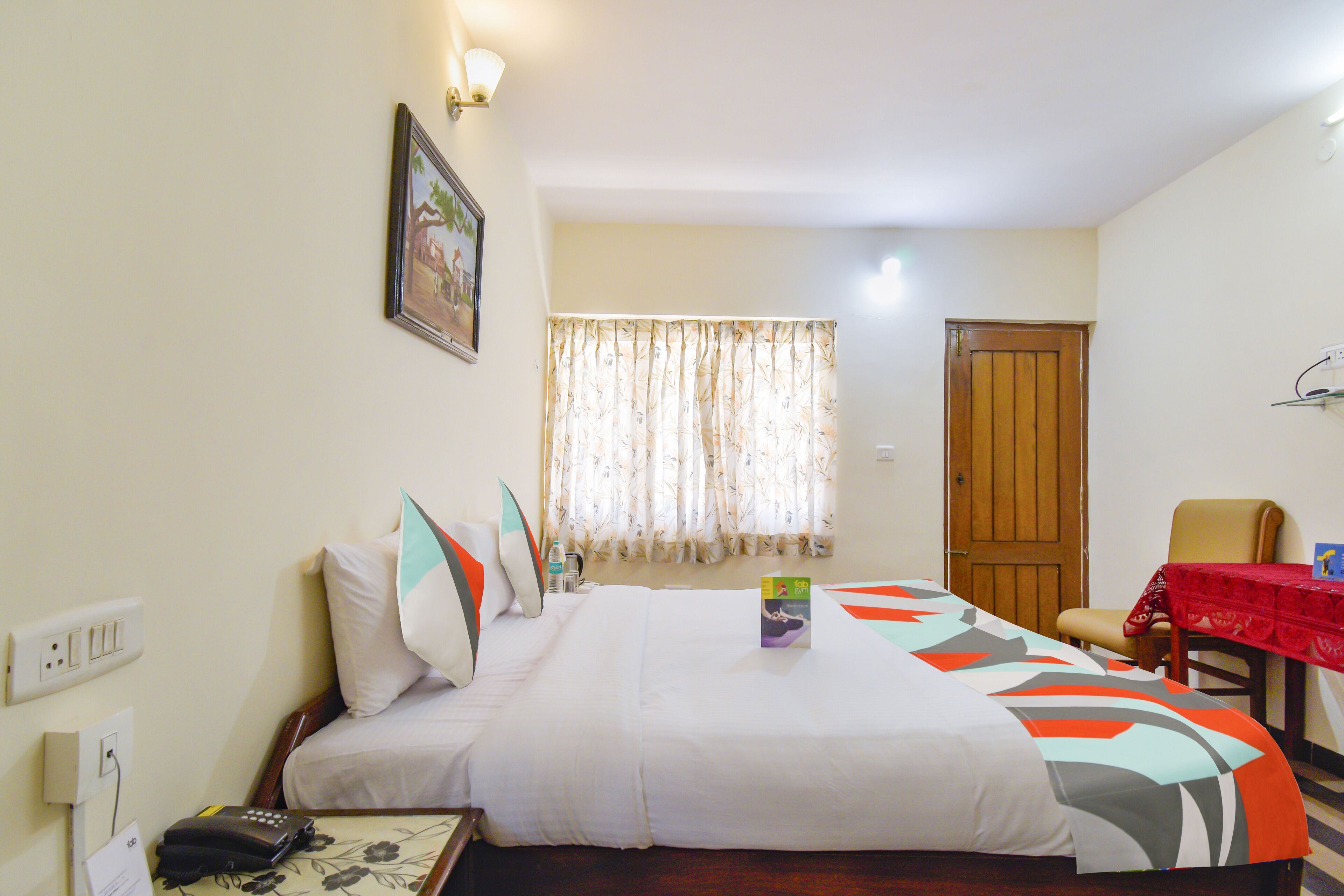 A hotel room with a bed, a chair, a table with a red tablecloth, a window with curtains, and a door.