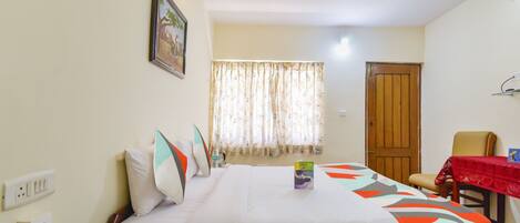 A hotel room with a bed, a chair, a table with a red tablecloth, a window with curtains, and a door.
