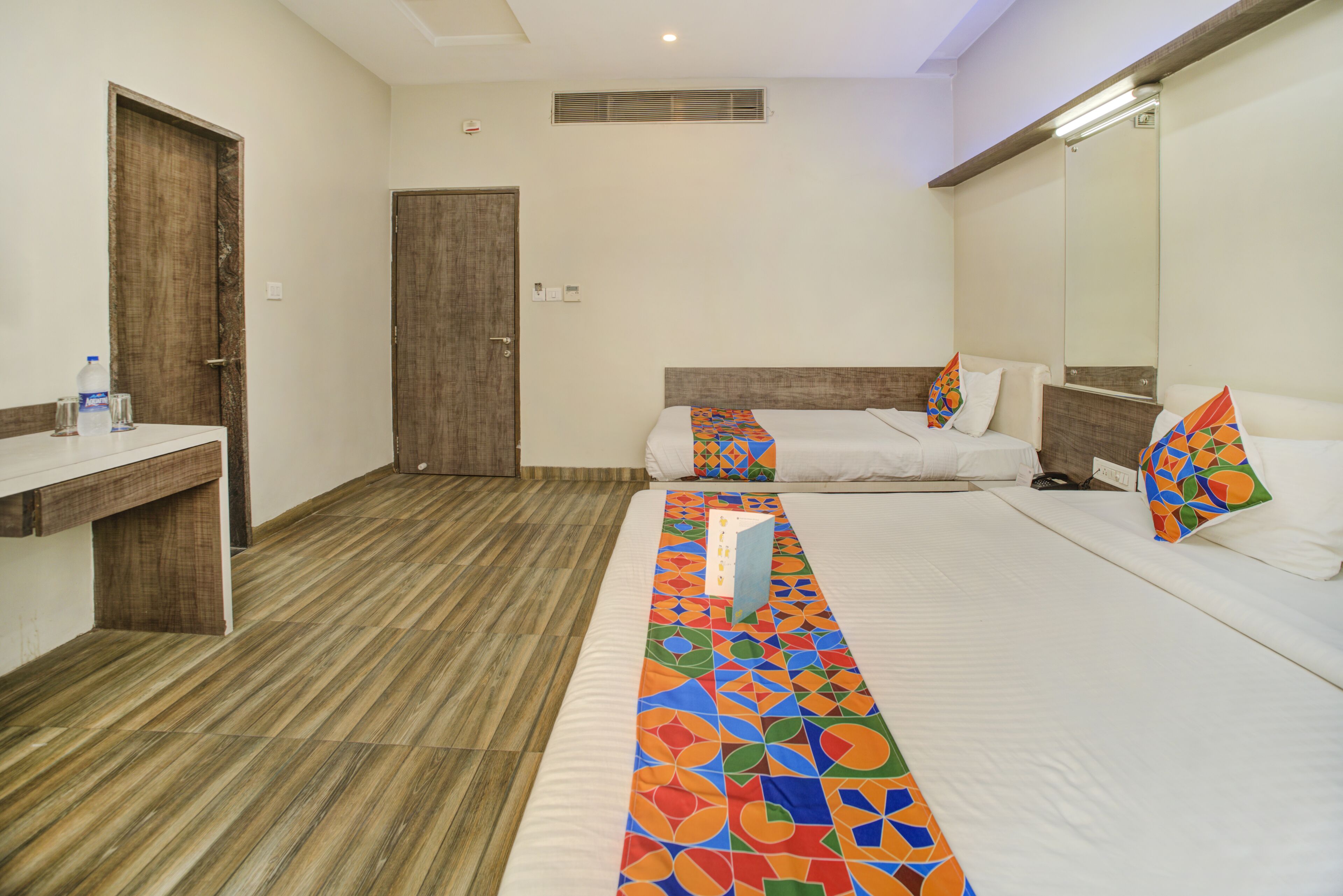 A hotel room with a wooden floor, a bed with a colorful geometric-patterned bedspread, a bedside table, a mirror, and a wooden door.