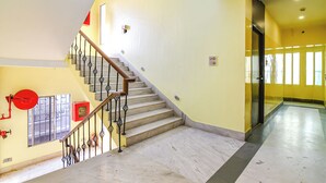 A staircase with a wooden handrail and black balusters, leading to a landing with a fire alarm and a window. - Fabhotel Aayash (Bidhannagar)