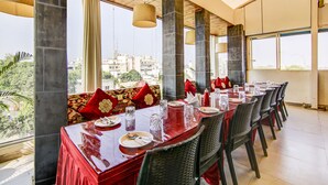Un espace repas avec une longue table dressée pour le repas, de grandes fenêtres offrant une vue sur la ville et de hauts plafonds avec des poutres apparentes.