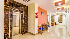 Un vestíbulo de hotel con un ascensor de mármol, un extintor, una zona de estar con sillas y una pared roja.
