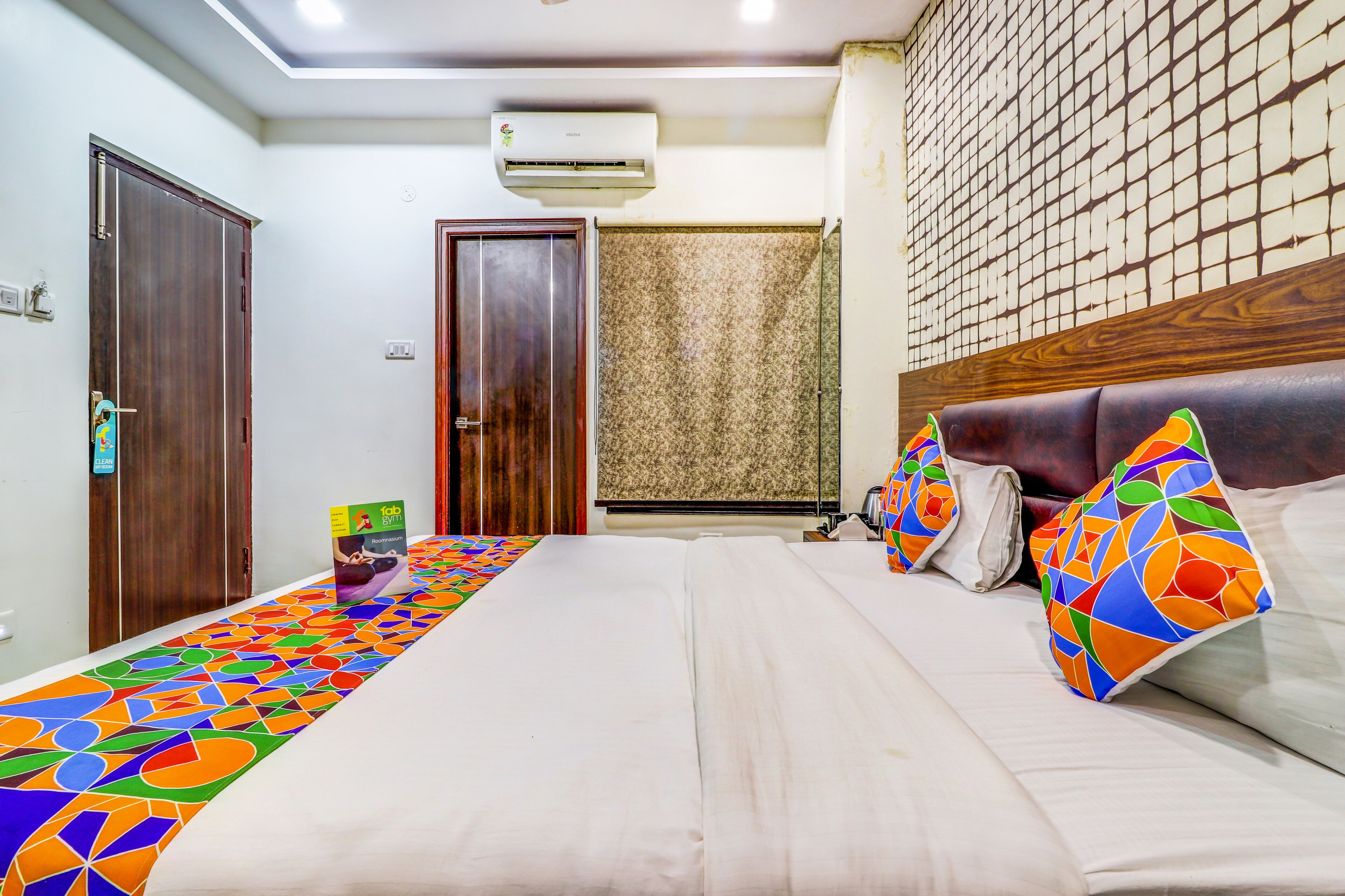 A hotel room with a bed, a headboard, a wall-mounted air conditioner, a patterned curtain, and a colorful bed runner.