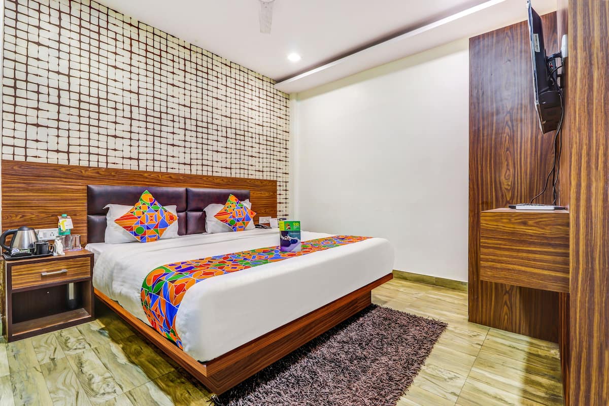 Habitación de hotel moderna con cabecera de madera, cama grande con ropa de cama colorida, mesita de noche con tetera y televisor montado en mueble de madera.