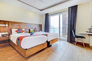 A hotel room with a wooden headboard, two beds with colorful bedding, a desk with a chair, and a large window with curtains. - Fabhotel Soft Petals (Gurugram)