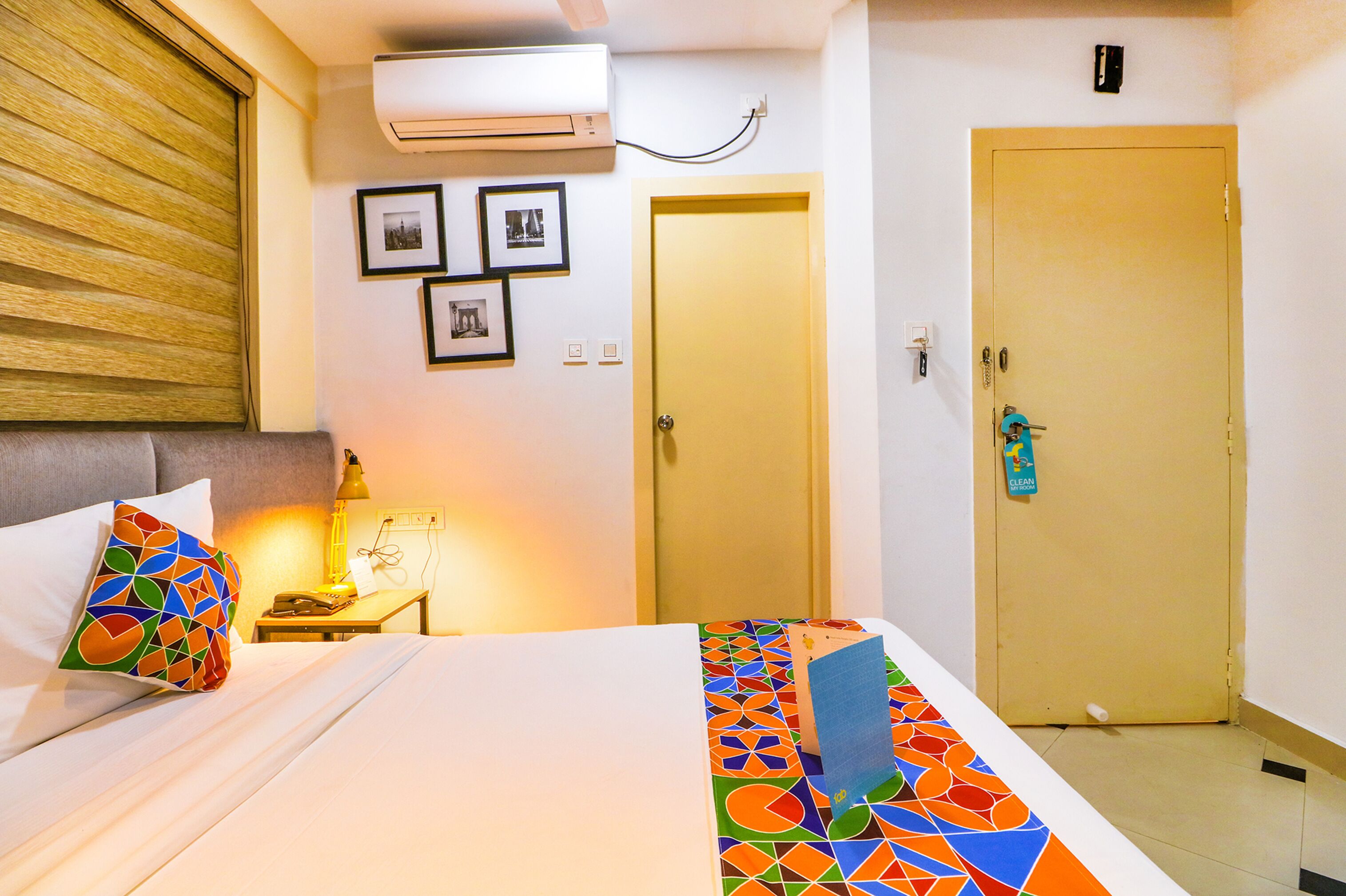 A hotel room with a bed, a nightstand, a lamp, an air conditioner, a door, and framed pictures on the wall.