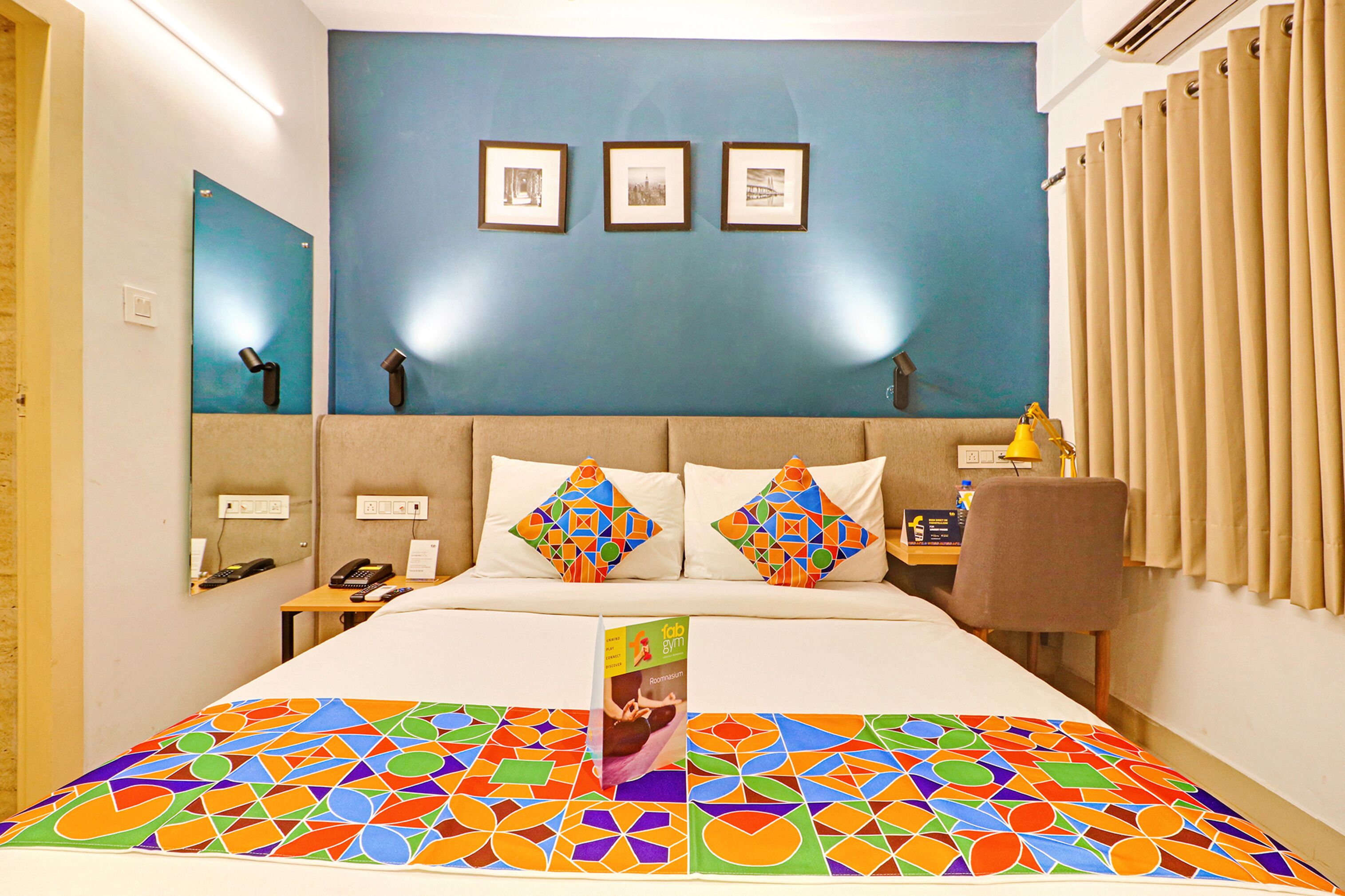A hotel room with a large bed, a desk, and a chair. There are three framed pictures on the wall and a mirror on the left wall.