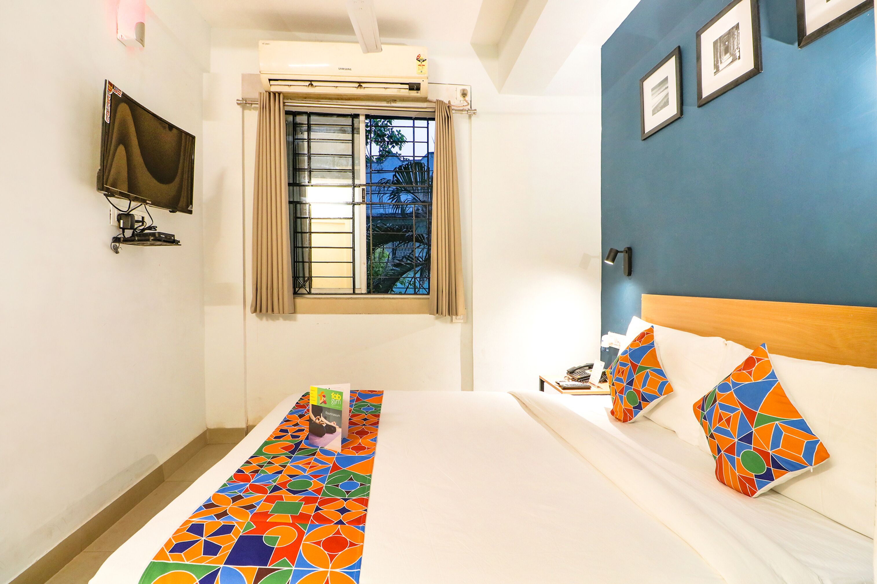 Une chambre d’hôtel avec un lit, un téléviseur fixé au mur, une fenêtre avec des rideaux et un mur bleu orné de tableaux.