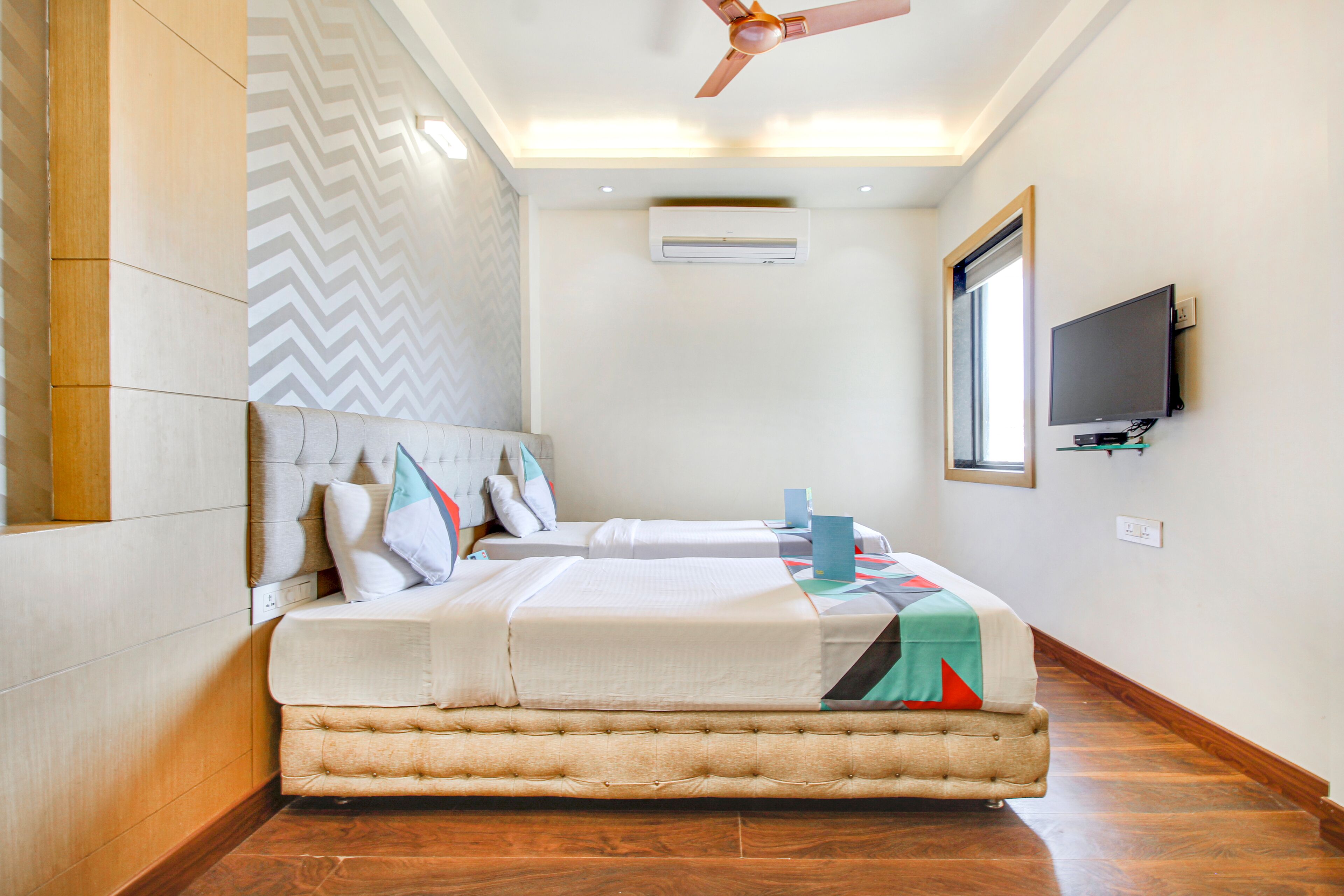 A hotel room with two beds, a ceiling fan, an air conditioning unit, a television, and a window.