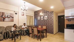 Una sala de estar con sofás estampados, una mesa de comedor con sillas, una araña y una zona de cocina con armarios.