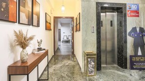 Un vestíbulo de hotel con un mostrador de recepción de madera, obras de arte enmarcadas, un jarrón con plantas secas, una mesa pequeña con un objeto decorativo, suelo de mármol, un ascensor y un cartel que dice 'NO CCTV EYEWATCHING'.