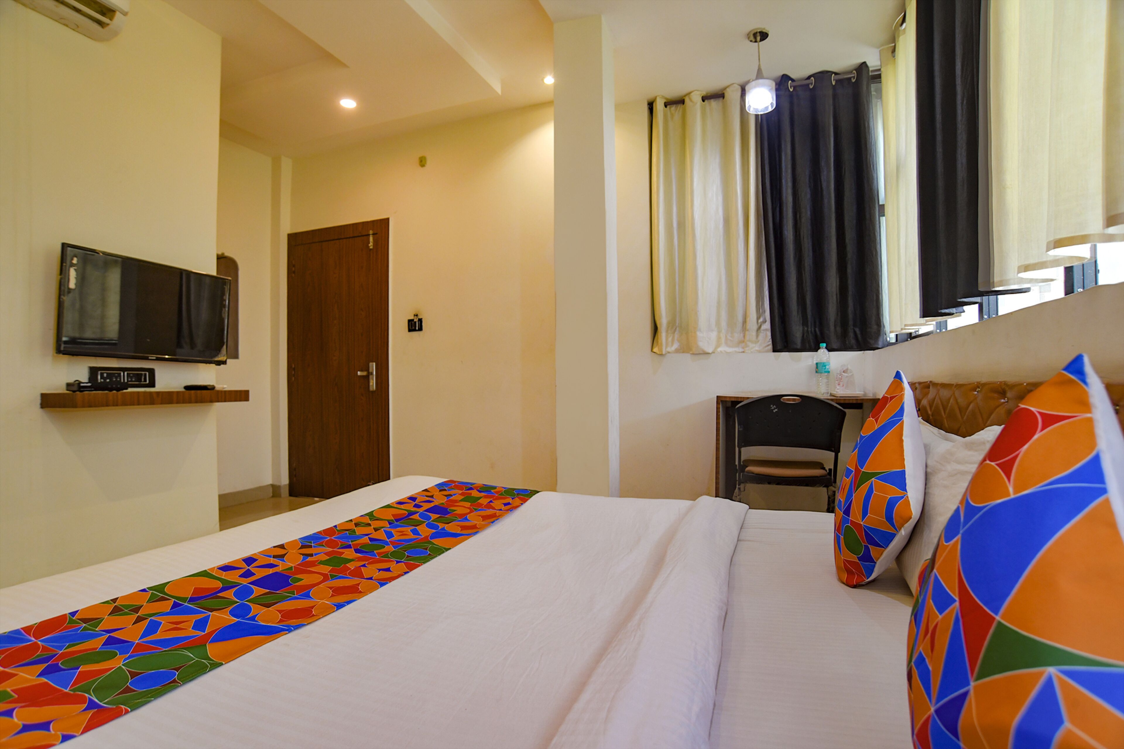 Une chambre d’hôtel avec un lit, un téléviseur fixé au mur, une fenêtre avec des rideaux et une petite table avec une chaise.