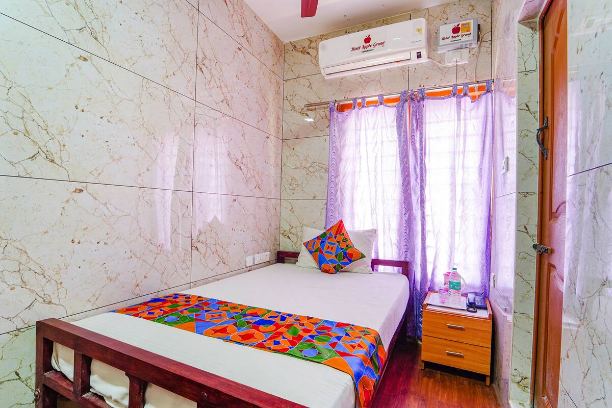 A hotel room with a bed, a bedside table with a bottle, and an air conditioning unit on the wall.