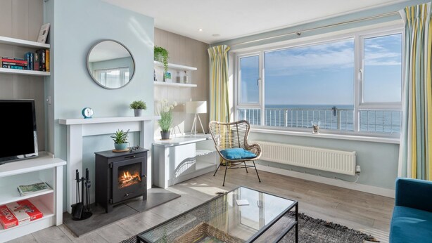 A modern living room with a fireplace, a television, and a view of the sea through large windows. - Dolphins Apartment - Spectacular Sea Views (Saundersfoot)