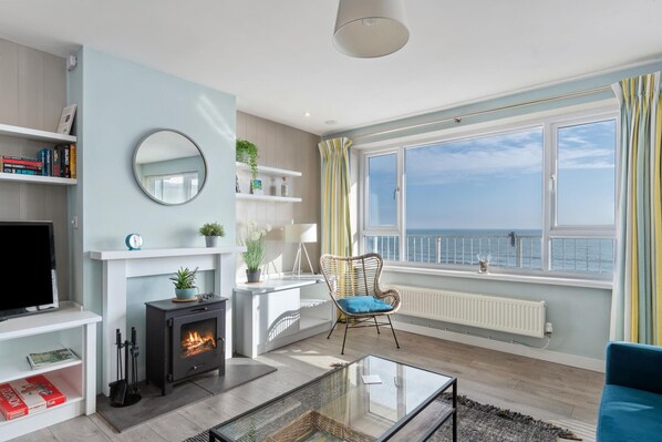 A modern living room with a fireplace, a television, and a view of the sea through large windows. - Dolphins Apartment - Spectacular Sea Views (Saundersfoot)