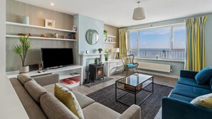 A modern living room with a flat-screen TV, sofa, and a blue armchair. There is a fireplace, a coffee table, and a view of the sea through large windows. - Dolphins Apartment - Spectacular Sea Views (Saundersfoot)