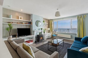 A modern living room with a flat-screen TV, sofa, and a blue armchair. There is a fireplace, a coffee table, and a view of the sea through large windows. - Dolphins Apartment - Spectacular Sea Views (Saundersfoot)