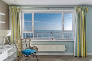 Ein Zimmer mit großem Fenster, einem Korbstuhl mit blauer Sitzkissen, einem weißen Heizkörper und einem weißen Schreibtisch mit Lampe.