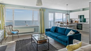 A modern living room with a blue sofa, a glass coffee table, and a view of the sea through large windows. - Dolphins Apartment - Spectacular Sea Views (Saundersfoot)
