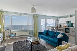 A modern living room with a blue sofa, a glass coffee table, and a view of the sea through large windows. - Dolphins Apartment - Spectacular Sea Views (Saundersfoot)