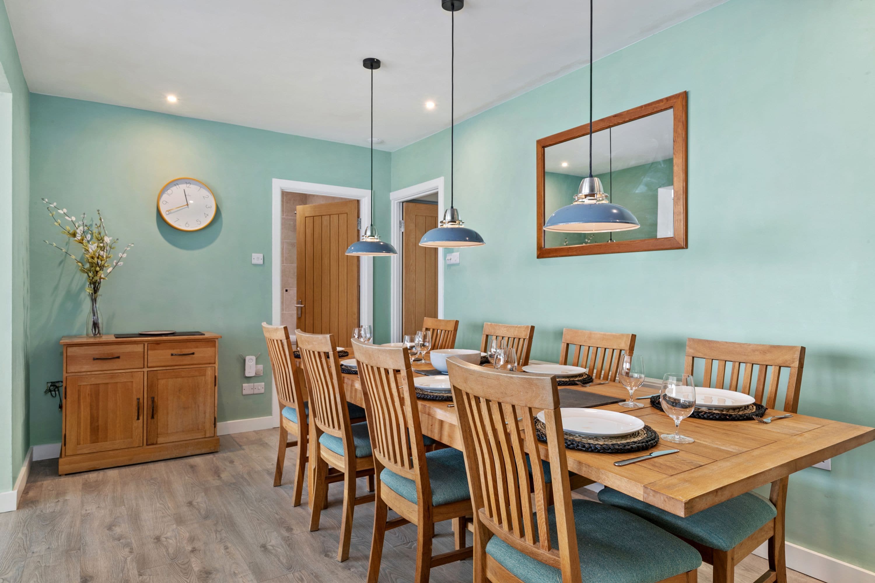 A dining room with a wooden table set for six, surrounded by six chairs with cushioned seats. There are two pendant lights hanging above the table, and a wooden cabinet to the left.