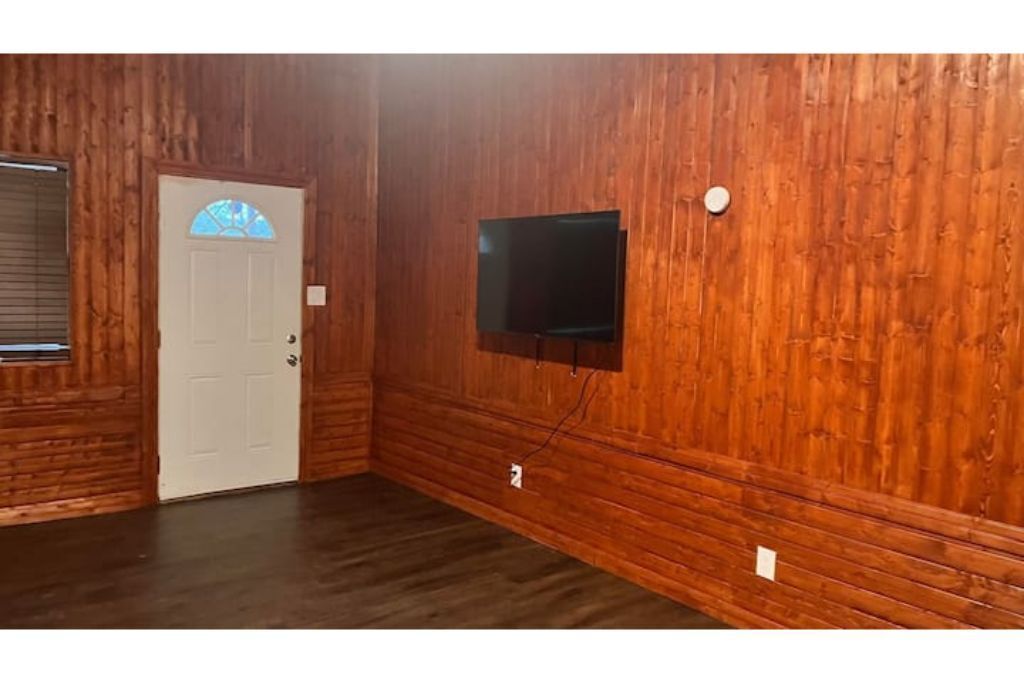 A room with wooden paneling, a white door with a blue window, and a mounted flat-screen TV.