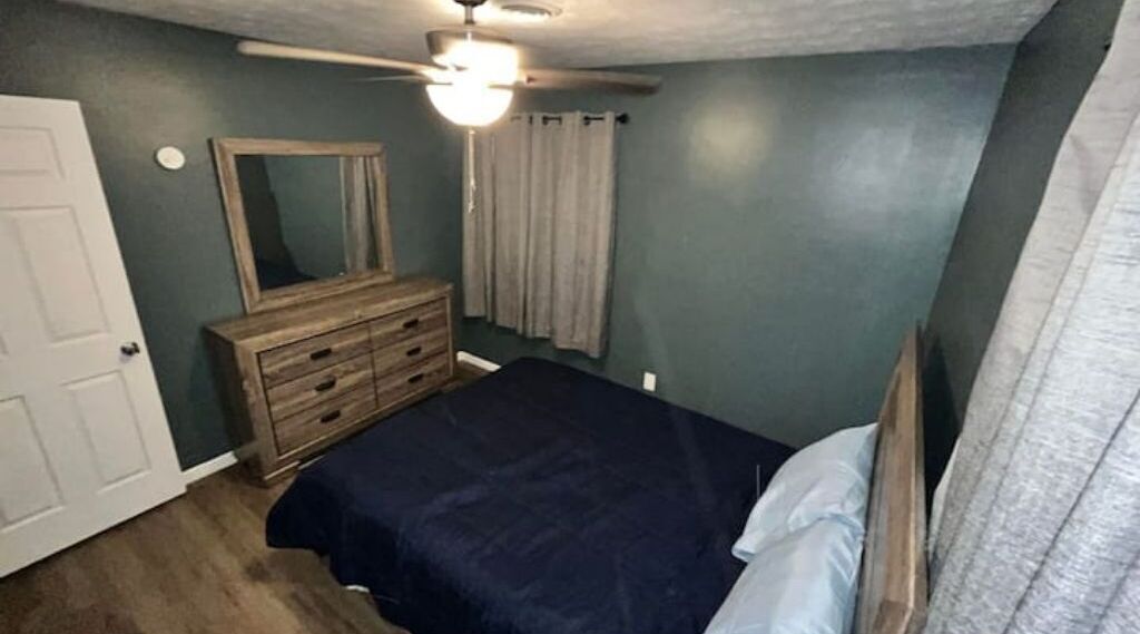Quarto com cama, penteadeira, ventoinha no teto e porta.