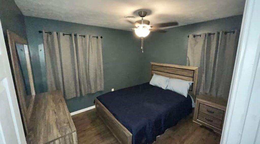 Un dormitorio con cama, mesita de noche, cómoda de madera, ventilador de techo y cortinas.